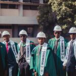 Group of chemical engineering graduates wearing academic gowns and hard hats outdoors.