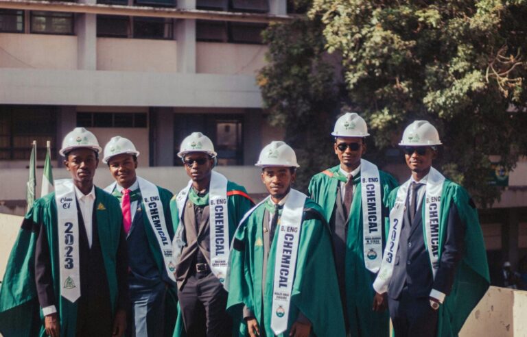 Group of chemical engineering graduates wearing academic gowns and hard hats outdoors.