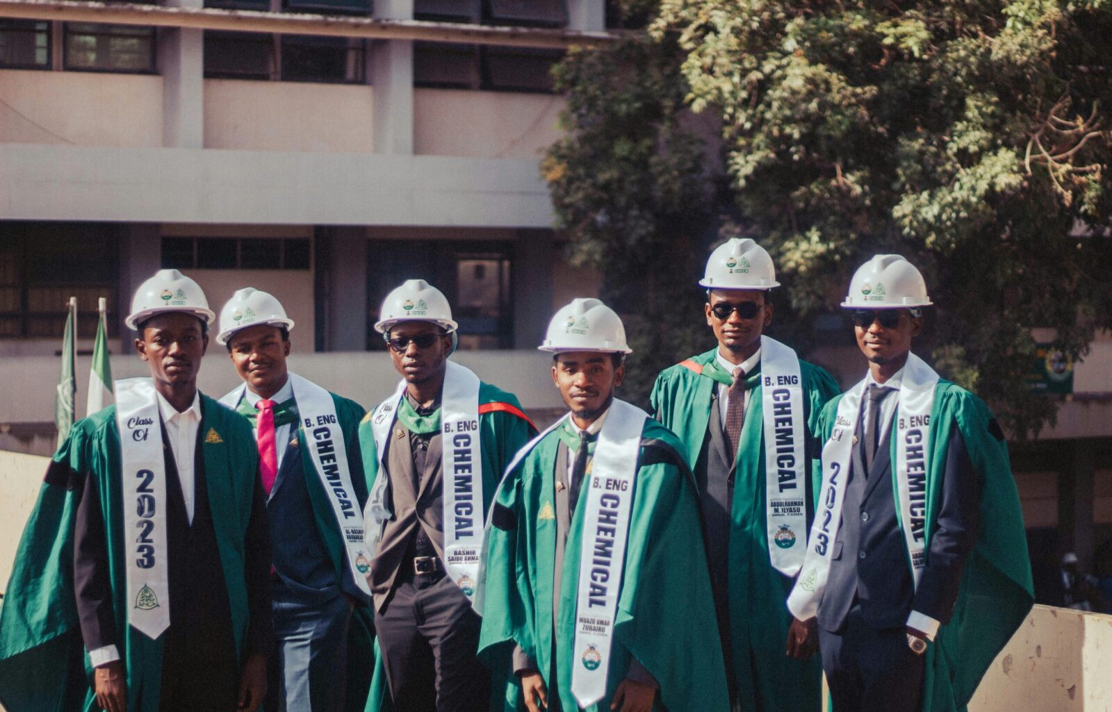Group of chemical engineering graduates wearing academic gowns and hard hats outdoors.