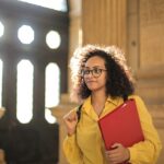 A young woman in a library carrying a red binder, bathed in sunlight.