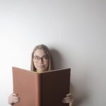 Positive young curious female wearing eyeglasses looking up while standing against gray wall with big opened book with brown cover in hands