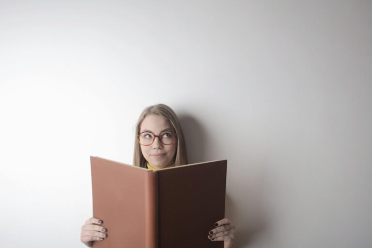 Positive young curious female wearing eyeglasses looking up while standing against gray wall with big opened book with brown cover in hands