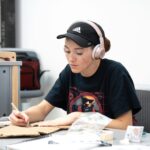 Young woman in headphones crafting at a table with cardboard for a school project, indoors.