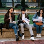 Three students studying together on a bench in a university campus setting outdoors.